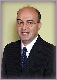 The image features a man wearing glasses, a suit, and a tie, with his head turned slightly towards the camera. He has dark hair and appears to be smiling. The background is neutral and does not provide any additional context about the setting or event.