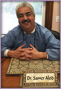 The image shows a man seated at a desk with a nameplate that reads  Dr. Samer Alois.  He is smiling, wearing glasses, and has his hands crossed on his lap. Behind him, there s a framed certificate on the wall above the desk.