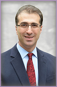 The image is a portrait of a man wearing glasses, with dark hair, dressed in a suit and tie, standing against a light background.