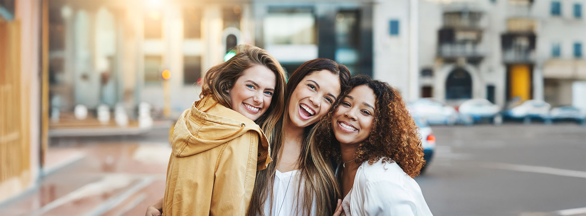 Four women posing together on a city street, smiling and happy, with a blurred background of an urban setting.