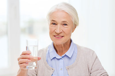 The image features an elderly woman holding a glass of water, smiling at the camera, with a clear background.