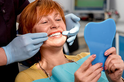 The image shows a woman sitting in a dental chair with her mouth open, receiving dental care  she holds a blue dental impression tray while smiling at the camera.