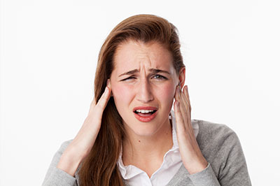 The image shows a young woman with her hand on her ear, possibly indicating discomfort or hearing difficulties, set against a plain background.