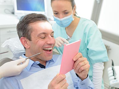 A man seated in a dental chair, holding up a pink card with a surprised expression, while a dentist stands behind him with a smile, holding a mirror.