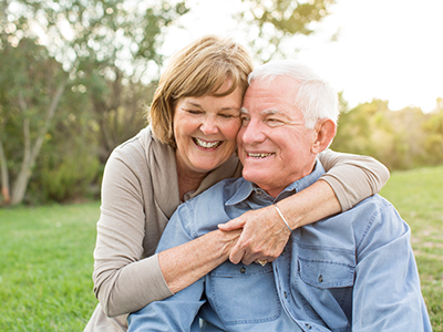 The image shows an elderly couple sharing a warm embrace, with the man standing behind the woman, both smiling and appearing content.