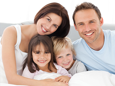 A family of four adults and two children posing together on a bed with a warm smile.