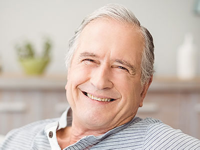 The image shows a man with gray hair, smiling and wearing glasses, sitting comfortably in a chair indoors.