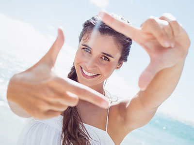 A woman with long hair is smiling at the camera while holding up her index finger, creating a frame with her hand. She is on a beach with clear skies in the background.